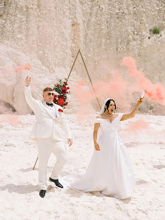 Couple portrait with wedding smoke bombs drifting pink as bride in off-shoulder gown and groom in tux pose by triangle arch on sand cliff