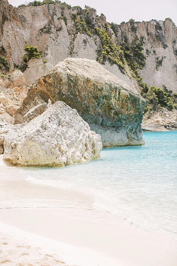 Beach scenery with turquoise water beach, white sand shoreline, and rock formations beneath coastal cliffs, trees, and open sky
