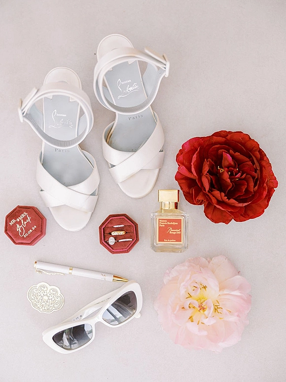 Bridal details flat lay with white bridal heels, engagement ring and band, perfume, sunglasses, and peonies on a neutral surface
