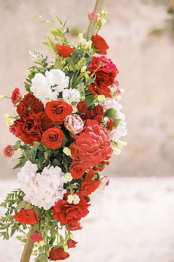 Wedding floral installation with red roses and white hydrangea on an asymmetrical arch frame against an outdoor sky backdrop