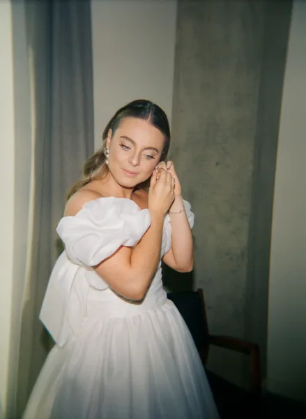 Bride getting ready, bride putting on earrings as she fastens pearl studs, soft waves framing her face in a classic white gown.