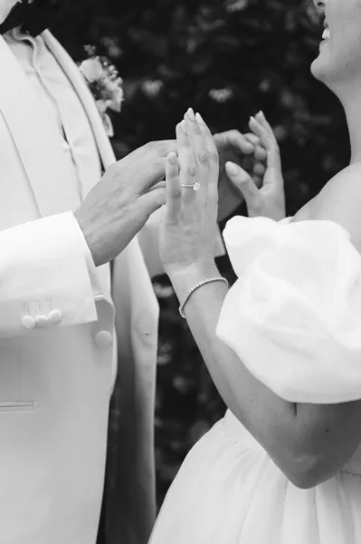 Wedding ring exchange, placing wedding band on the groom's finger as her puff sleeves brush close, set against lush garden greenery.