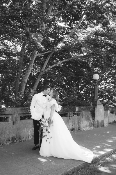 Couple portrait capturing a wedding kiss moment as the bride holds a cascading bouquet beneath trees by a stone railing and lamppost.