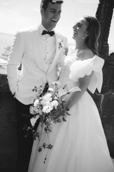 Couple portrait of a bride and groom laughing by a stone wall beside the water, she holds a white and green bridal bouquet.