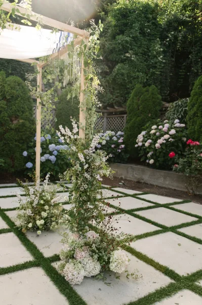 Wedding chuppah, a greenery chuppah under sunlight, stands before garden hedges and a lattice fence on concrete pavers.