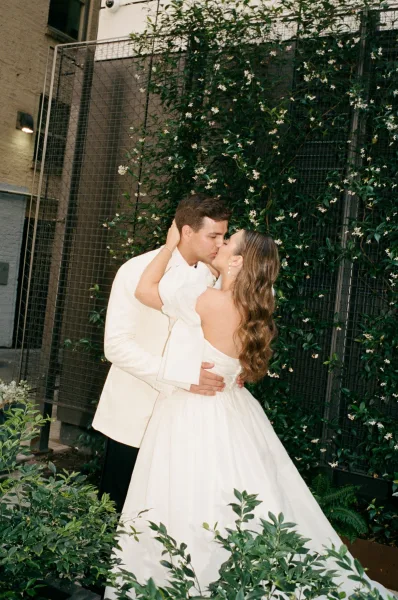 Wedding kiss portrait as the bride and groom kiss against a flowering vine trellis in a courtyard, her bow-detail gown.