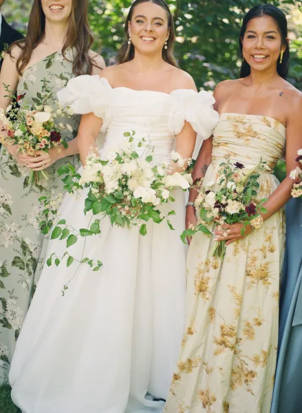 Bride and bridesmaids in mismatched bridesmaid dresses stand with bouquets amid garden greenery and trees, sharing a joyful moment.