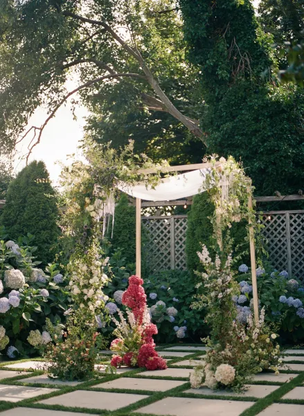 Ceremony arch styled as a wedding canopy with draped fabric, set among garden trees, hydrangea bushes, and a lattice fence.