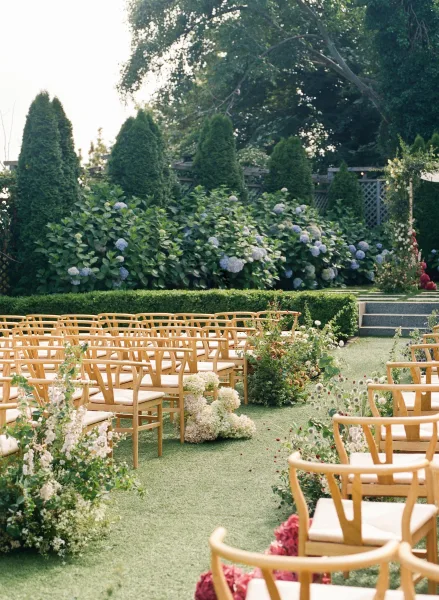 Ceremony setup with outdoor ceremony seating, hydrangea aisle flowers on the lawn leading toward a boxwood hedge and lattice fence backdrop.