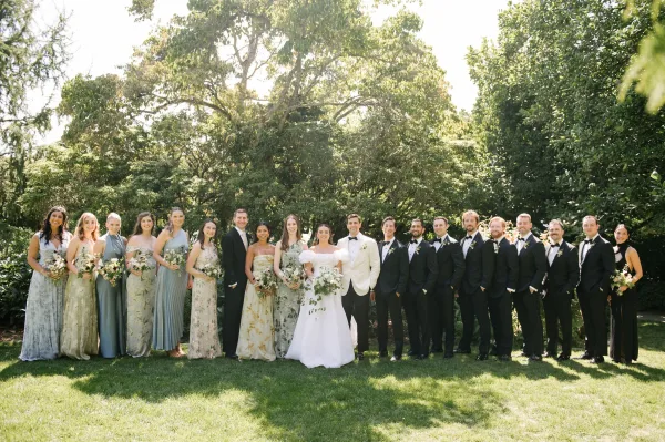 Wedding party portrait—a large bridal party photo—on a sunlit garden lawn, bridesmaids holding bouquets with the couple centered.