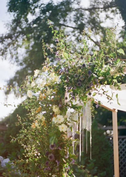 Floral wedding arch, a garden ceremony arch adorned with draped fabric, set against garden trees and a lattice fence.