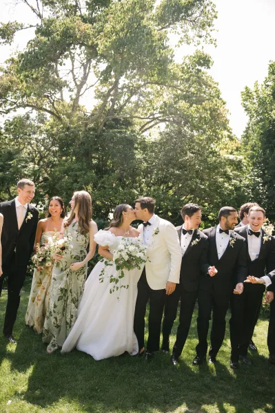 Wedding party portrait of newlyweds with wedding party under garden trees on a grass lawn, the bride holding a bridal bouquet.
