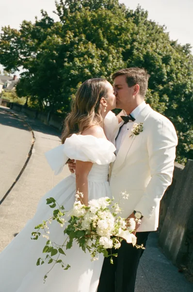 Wedding kiss portrait as bride and groom kiss, bride cradling a bridal bouquet on a tree-lined street beside a low stone wall.