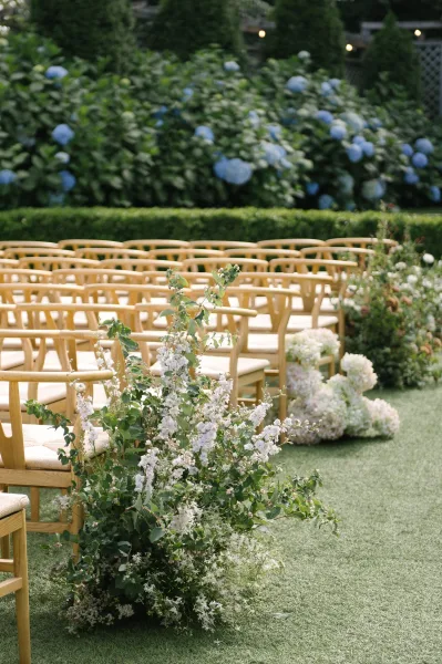Ceremony aisle design in outdoor ceremony decor, white hydrangea clusters edge the path amid garden hedges and a lattice fence.