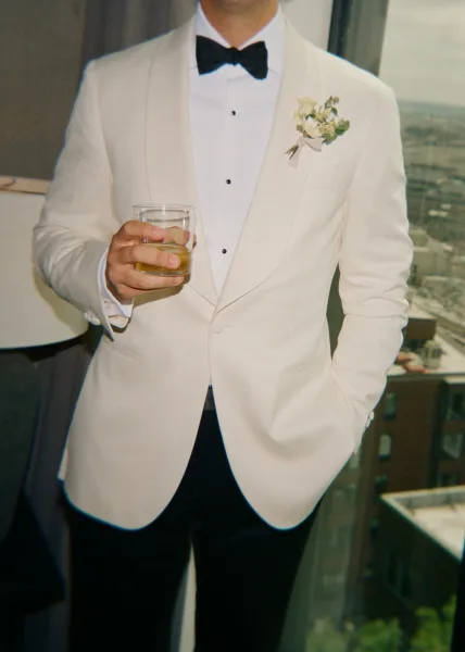 Groom portrait in an ivory dinner jacket, holding a whiskey glass by a window with a soft city skyline backdrop