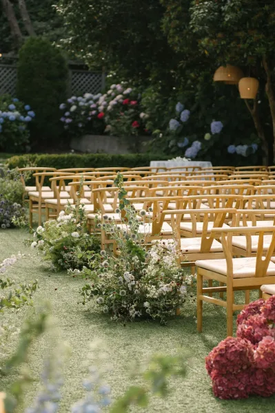 Ceremony aisle design with outdoor ceremony seating, a flower-lined path beneath rattan pendant lights, set amid hydrangeas and garden trees.
