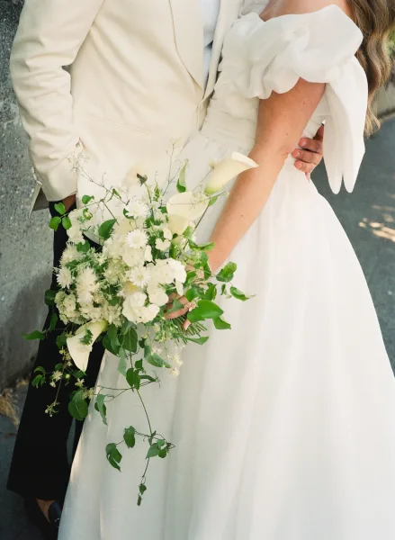 Bridal bouquet, a white and green bouquet with calla lilies resting by a stone wall on an outdoor walkway in dappled light.