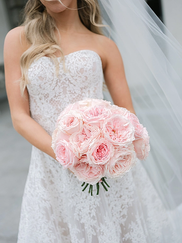 Bridal bouquet of pink garden roses held by a bride in a strapless lace wedding dress and veil on an outdoor walkway with soft backdrop