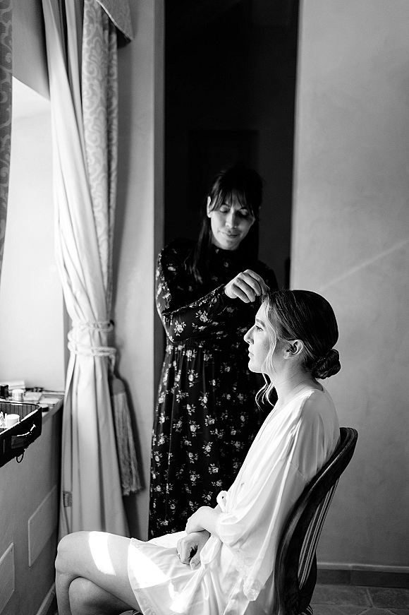 Bridal hair styling as a hairstylist shapes a low bun updo on the bride in a bridal robe by window light in an interior room