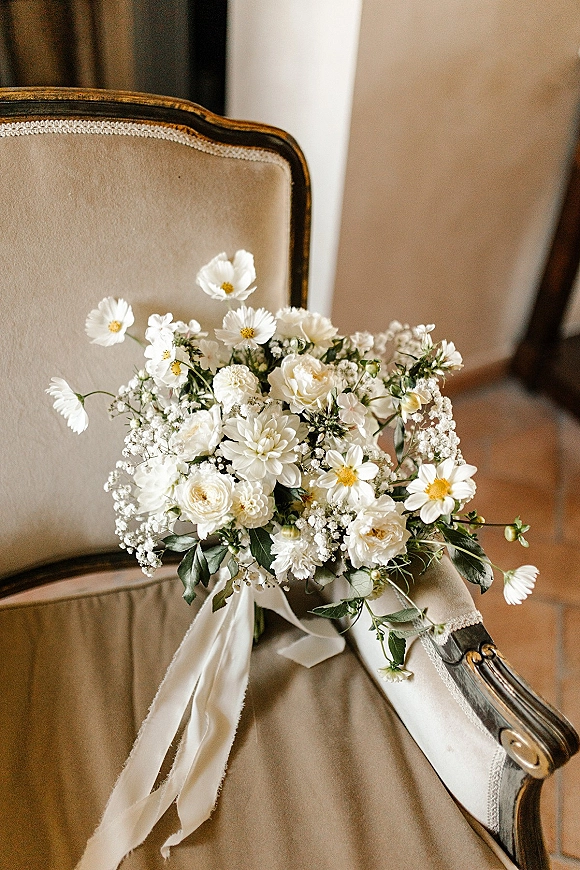 Bridal bouquet of white flowers and greenery with baby's breath and a silk ribbon draped over a vintage upholstered chair indoors