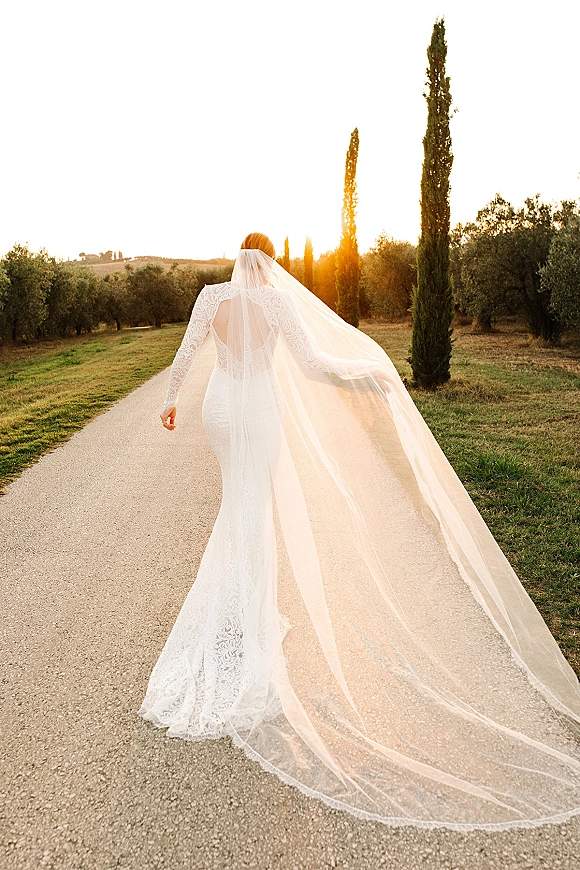 Bridal portrait of a bride walking away in a lace long sleeve wedding dress with cathedral veil on a country road at sunset