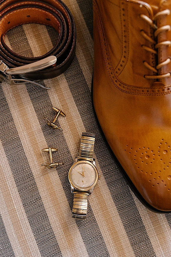 Groom accessories flatlay featuring tan leather dress shoes, belt, gold cufflinks and wristwatch arranged on a striped fabric surface