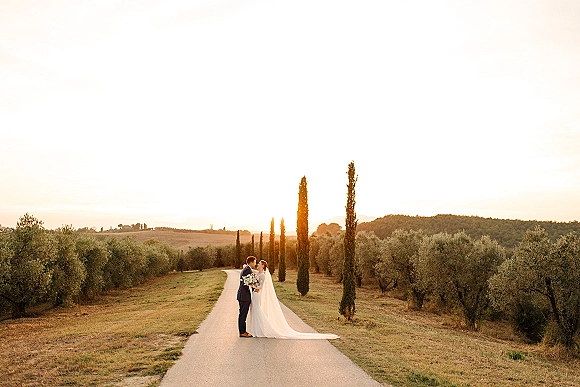 Couple portrait of bride and groom kiss on a country road at sunset, veil blowing, bouquet in hand, with cypress and olive hills behind