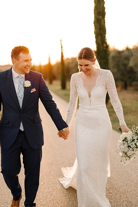 Couple portrait of bride and groom walking hand in hand on a tree-lined path at sunset, bride in lace sleeves with veil and bouquet