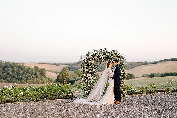 Wedding couple portrait of bride and groom embracing under a floral arch, her lace dress and veil framed by rolling countryside hills