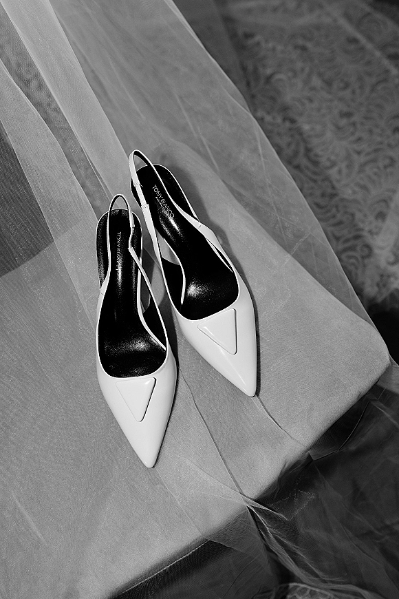 Bridal shoes, white bridal heels in pointed-toe slingback style resting on a sheer tulle veil beside an upholstered chair on patterned carpet