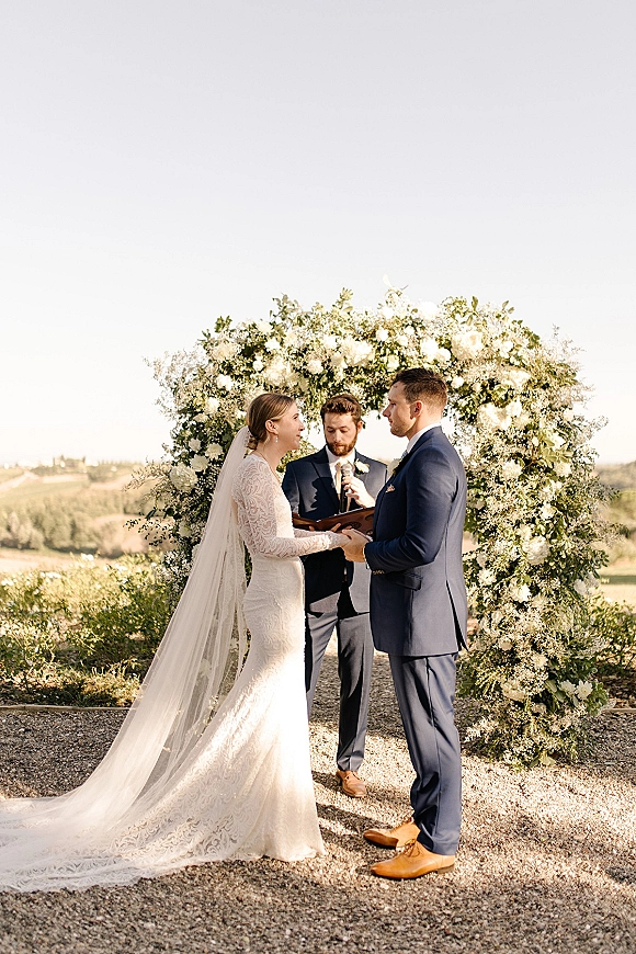 Wedding vows as bride and groom hold hands under a floral ceremony arch, officiant with microphone before hills and open sky