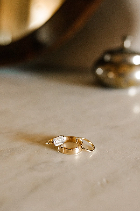 Wedding rings in a three ring stack, featuring gold wedding bands and an emerald cut engagement ring on a marble surface with blur behind