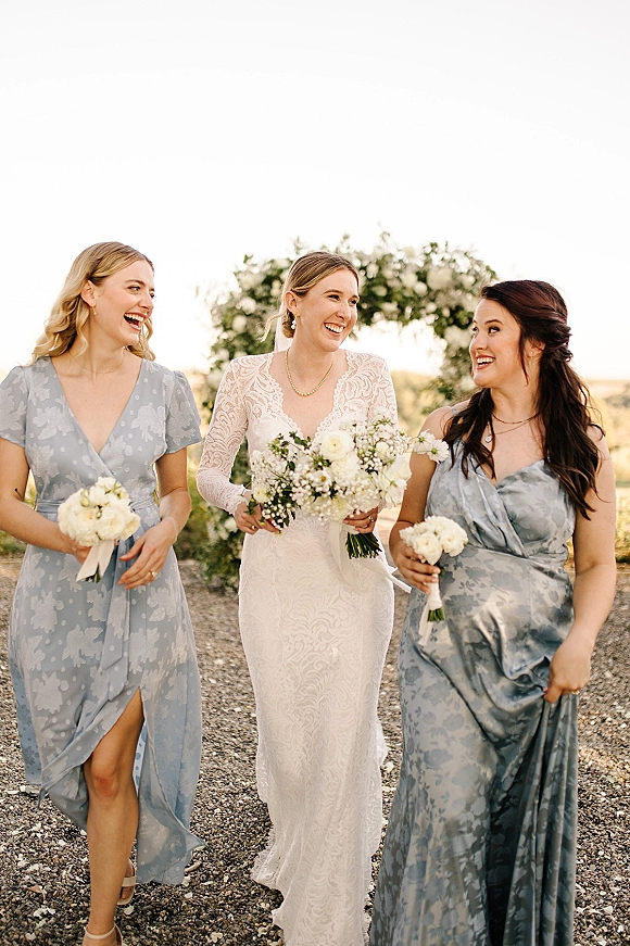 Bride with bridesmaids walking along a gravel path, holding white wedding bouquet, with dusty blue dresses and greenery landscape backdrop