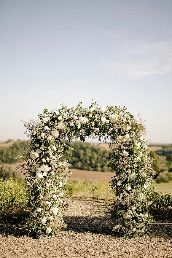 Wedding ceremony arch with white flowers and greenery, set on a gravel path with hillside fields, trees, and open sky beyond