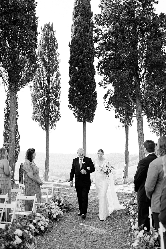 Wedding processional as bride walking down aisle with her father, holding bouquet and veil, past white chairs and aisle florals outdoors
