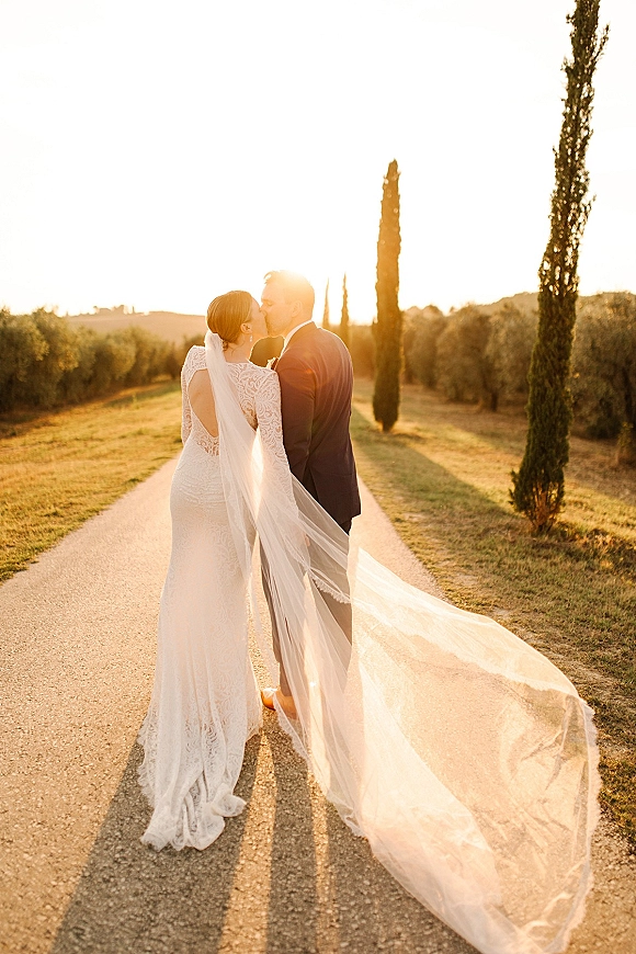 Wedding kiss portrait of bride and groom kissing, lace dress and long veil backlit by sunset on a country road lined with cypress trees