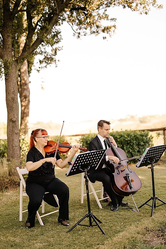 Wedding ceremony musicians, a string duo wedding playing violin and cello beside music stands on a lawn with trees and a wooden fence