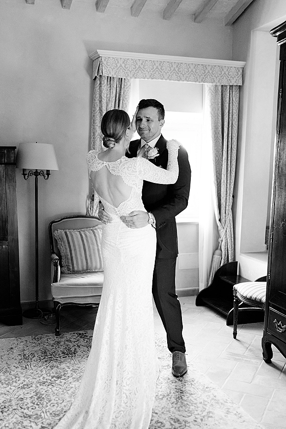 First dance as bride in a lace long sleeve open-back gown hugs groom in suit, lit by window light beside curtains and armchair indoors