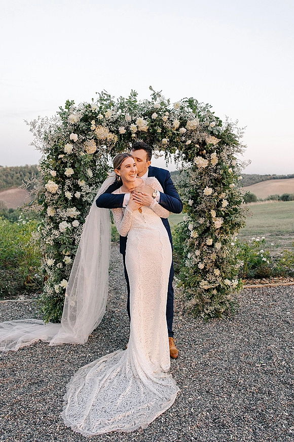 Couple portrait of bride and groom embrace as he kisses her forehead beneath a white floral arch, her long lace veil trailing outdoors