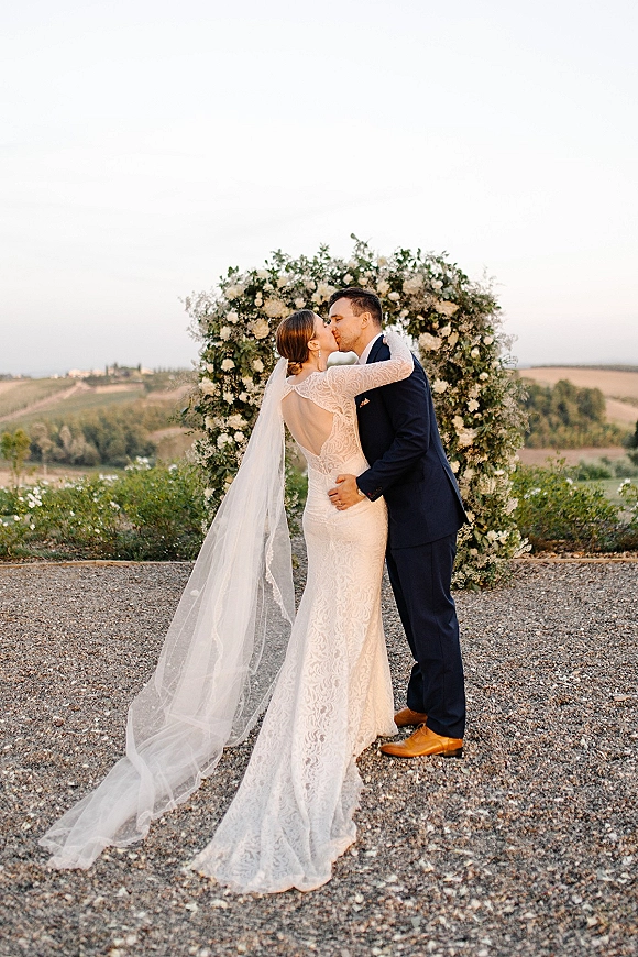 Wedding kiss portrait of bride and groom kissing beneath a circular floral arch, her long veil flowing against a hillside sky backdrop