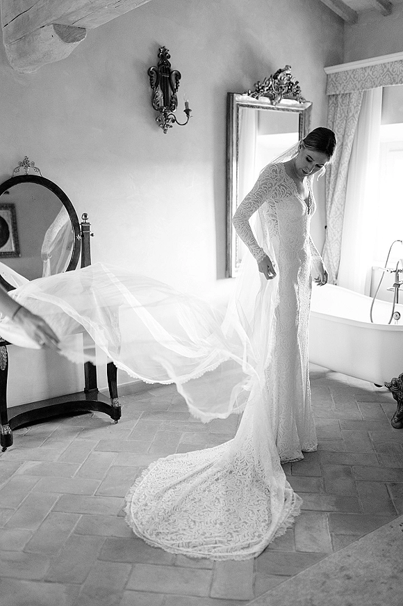 Bride getting ready in a lace wedding dress with long sleeves and veil, posing by a mirror near a freestanding bathtub in soft window light