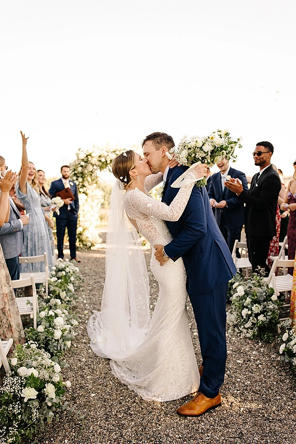 Wedding kiss as the couple shares a first kiss wedding moment on a gravel aisle, bride in lace sleeves and veil holding roses under open sky