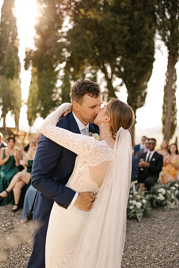 Wedding kiss as newlyweds embrace on a gravel aisle, bride in long-sleeve lace gown and veil, guests and trees glowing in sunlight