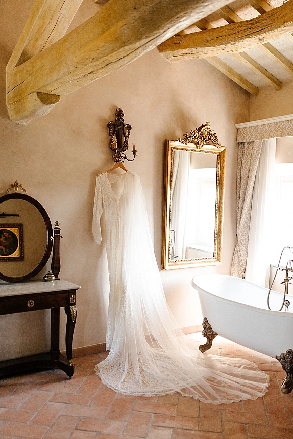 Wedding dress with lace long sleeves hanging from a wooden hanger beside an ornate gold mirror in a vintage suite with window light