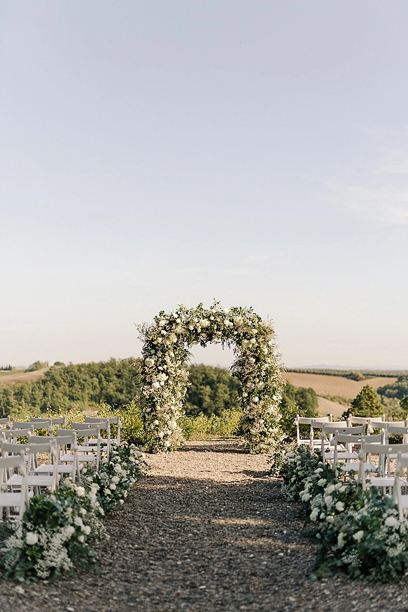 Ceremony setup for an outdoor wedding ceremony with a floral wedding arch, white chairs, and aisle flowers overlooking rolling hills and fields