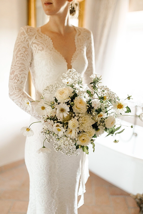 Bridal bouquet of white wedding bouquet roses, daisies, and baby’s breath with greenery held by bride in lace sleeves by window light