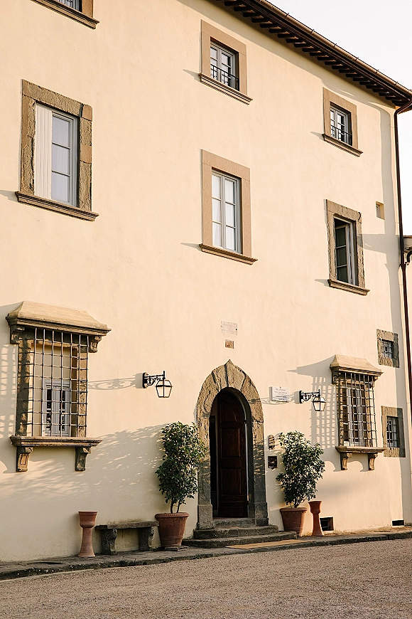 Wedding venue exterior of a cream stucco villa with a stone arch entrance, lanterns, potted trees, and iron-grilled windows under eaves