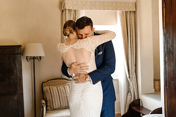 First look moment as bride in a lace wedding dress taps groom’s shoulder in a hotel room, soft window light and curtains behind