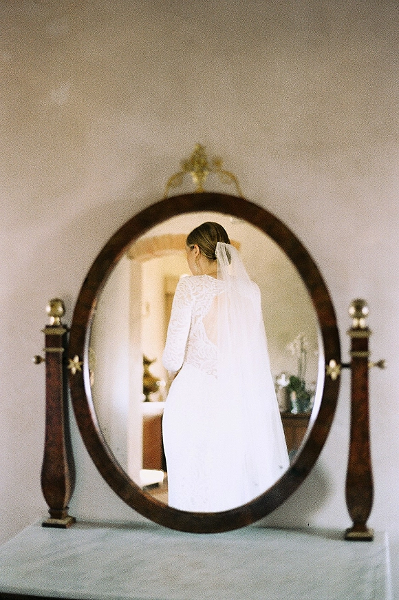 Bridal portrait of a bride in mirror, seen from behind in a lace-sleeved wedding dress and veil, reflected in an oval wooden frame indoors