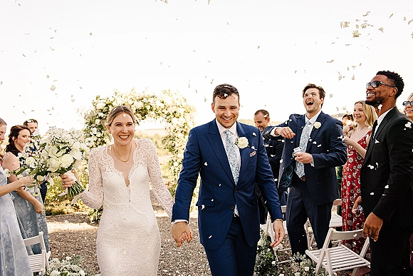 Wedding recessional as bride and groom walking aisle hand in hand under a floral arch, petals confetti flying on a gravel path outdoors